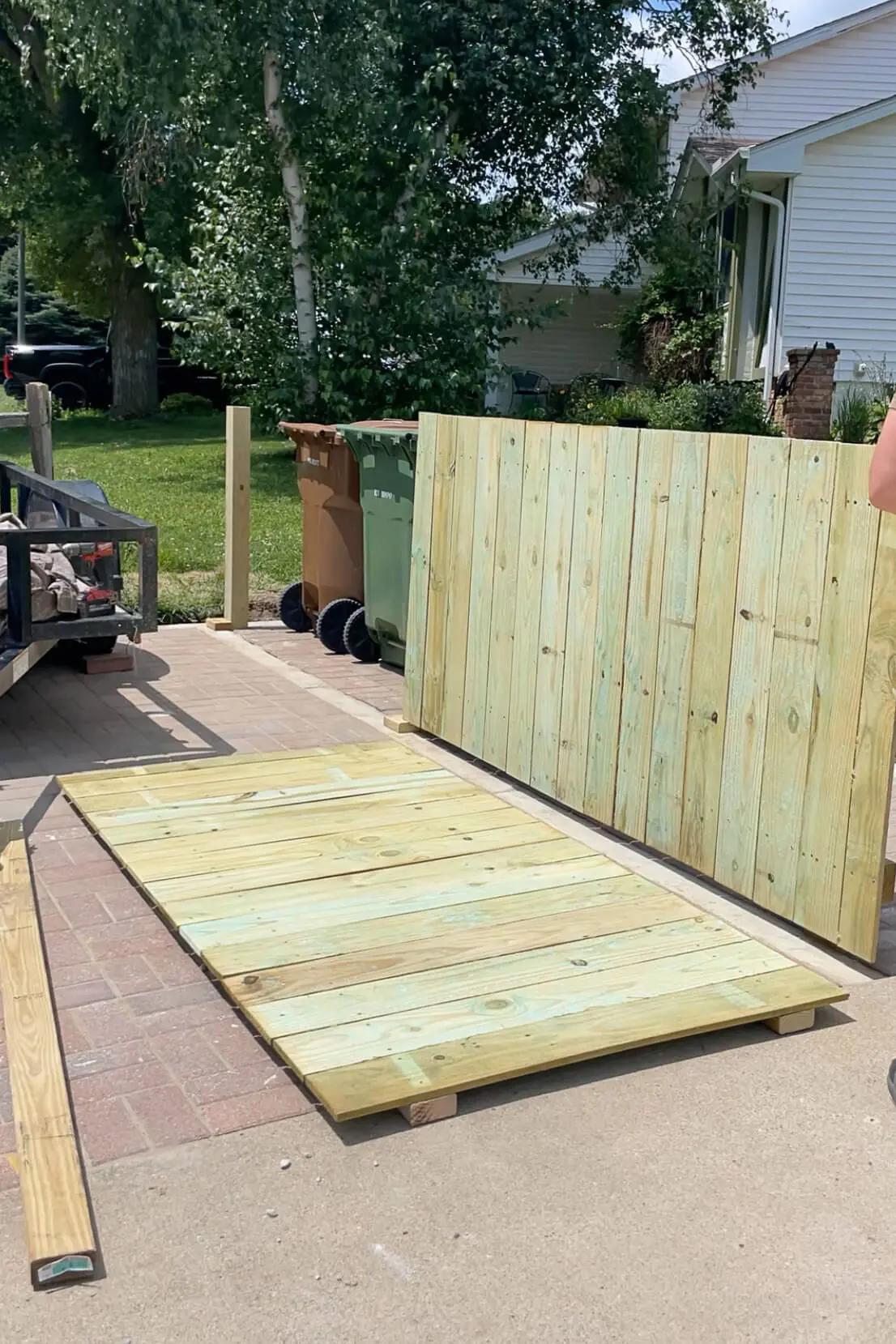 Two completed trash enclosure panels made from pressure-treated wood, laid out on a paver surface with trash bins staged nearby.

